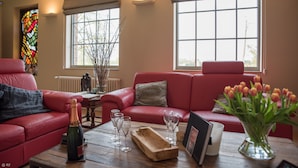 Un salon avec un canapé en cuir rouge, une table basse en bois sur laquelle se trouvent une bouteille de champagne et des verres, un vase de tulipes et un vitrail.