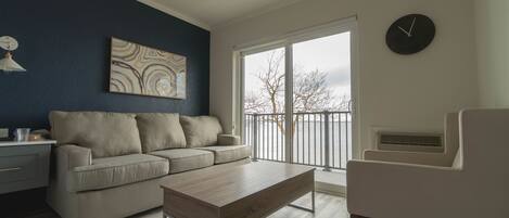 Sala de estar con sofá, mesa de centro y una silla. Se ve una ventana con vistas a árboles y un reloj en la pared.