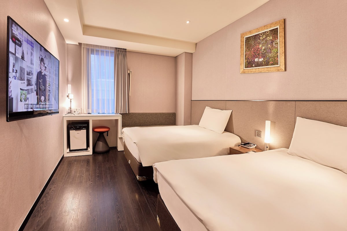 A hotel room with two beds, a desk with a chair, a television, and a painting on the wall.