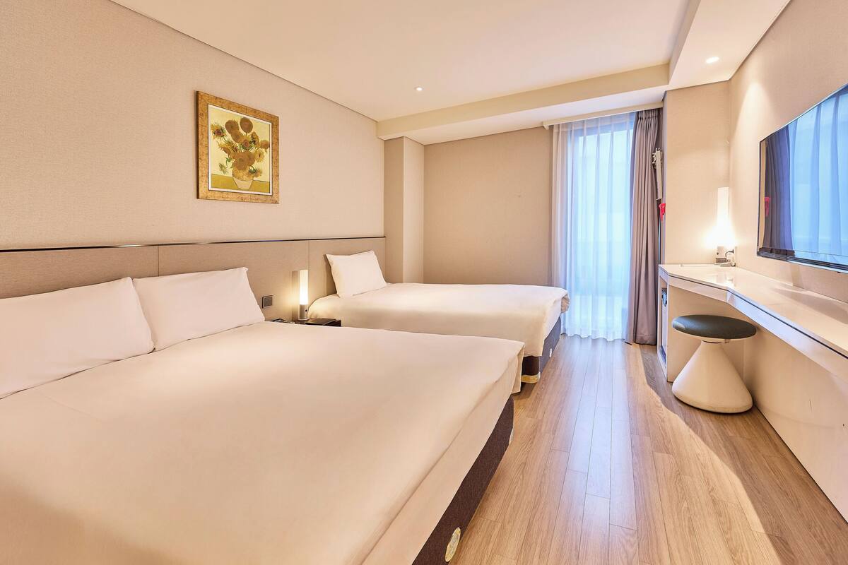 A hotel room with two beds, a bedside table with a lamp, a large mirror, and a window with curtains.