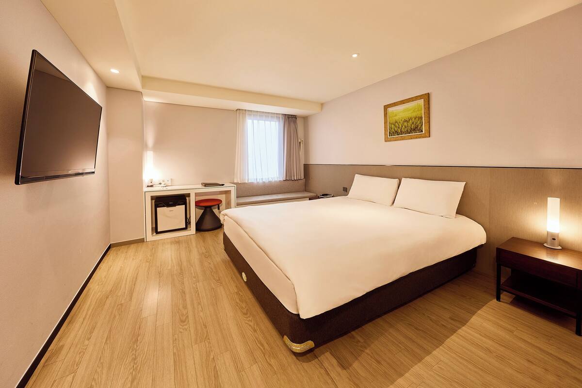 A hotel room with a large bed, a desk with a chair, a flat-screen TV mounted on the wall, and a bedside table with a lamp.