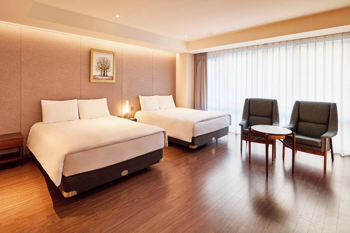 A hotel room with two beds, a chair, a small table, and a large window with curtains.