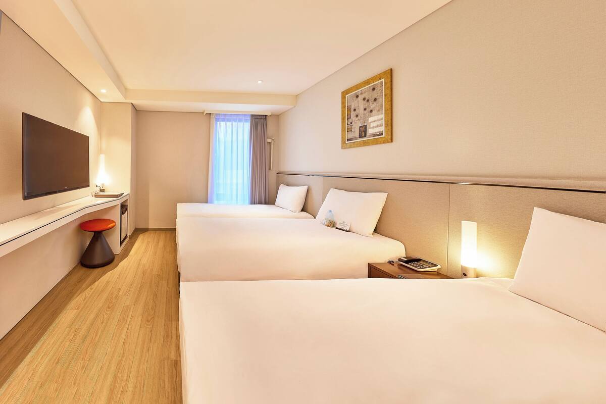 A hotel room with two beds, a TV, a bedside table with a lamp, and a window with curtains.
