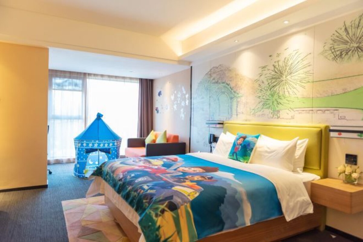 Una habitación de hotel con una cama grande, una mesita de noche, una carpa de juegos azul y un mural en la pared.
