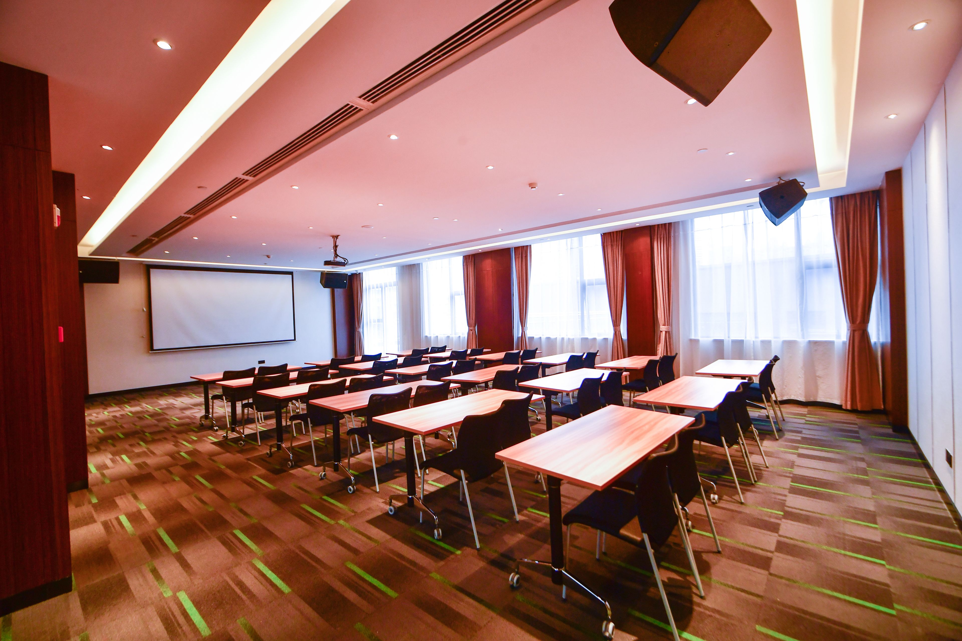 A conference room with multiple tables and chairs, a projector screen, and large windows with curtains.