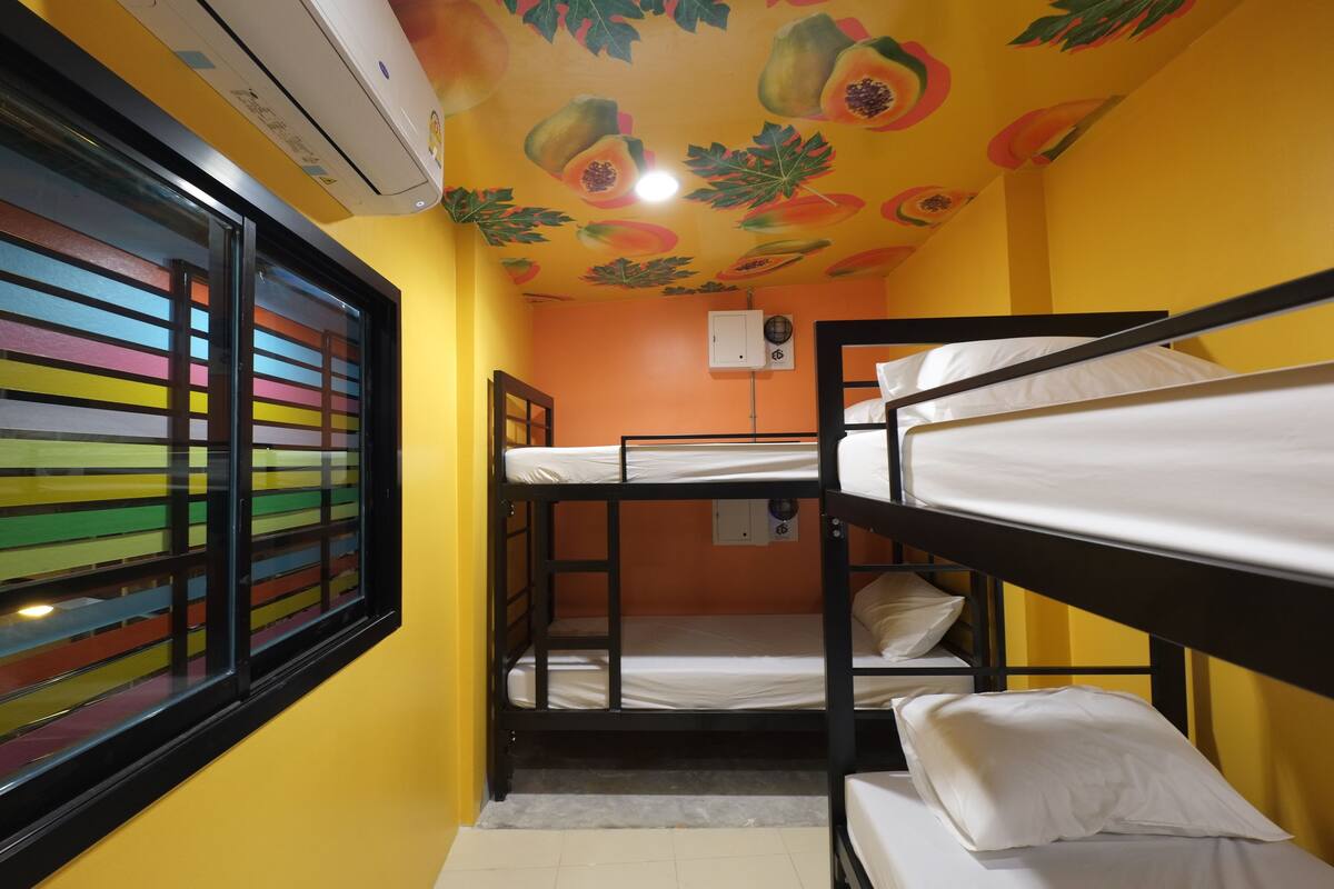 Une chambre de dortoir avec des lits superposés, une fenêtre avec des stores colorés et un plafond décoré de motifs de fruits.