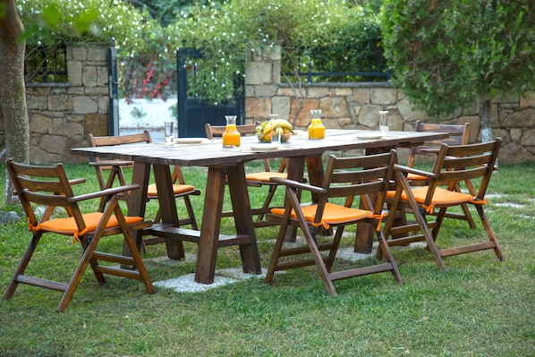 Une table en bois, entourée de chaises, de verdure et d’un mur en pierre.