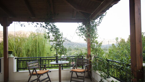 A covered outdoor seating area with two wooden chairs and a small table, overlooking a lush garden and distant mountains. - Villa Jasmine (Agios Vasileios)