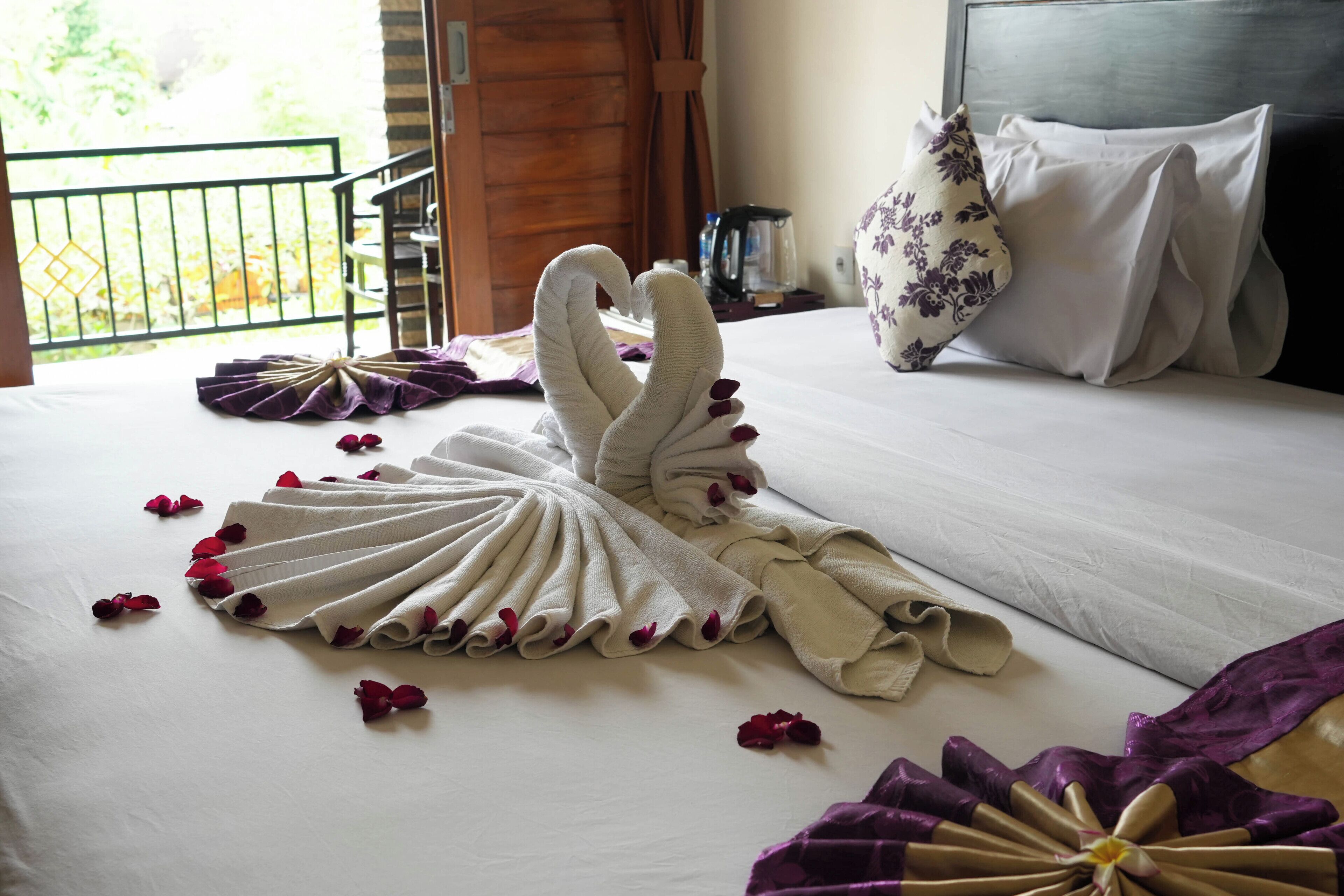 A neatly made bed with a towel arranged in a swan shape, scattered rose petals, and a purple and gold decorative item.