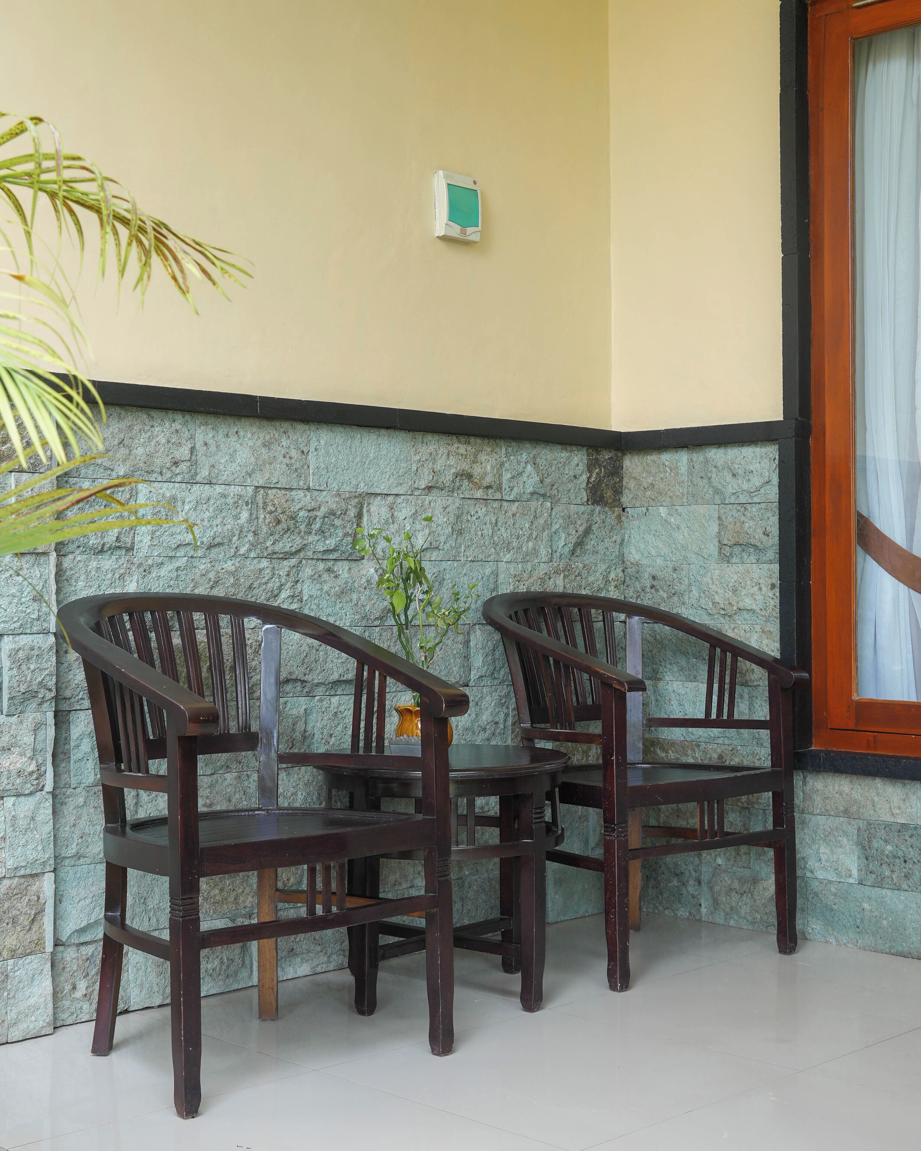 A corner of a room with two wooden chairs and a small table, a stone wall, and a window with curtains.