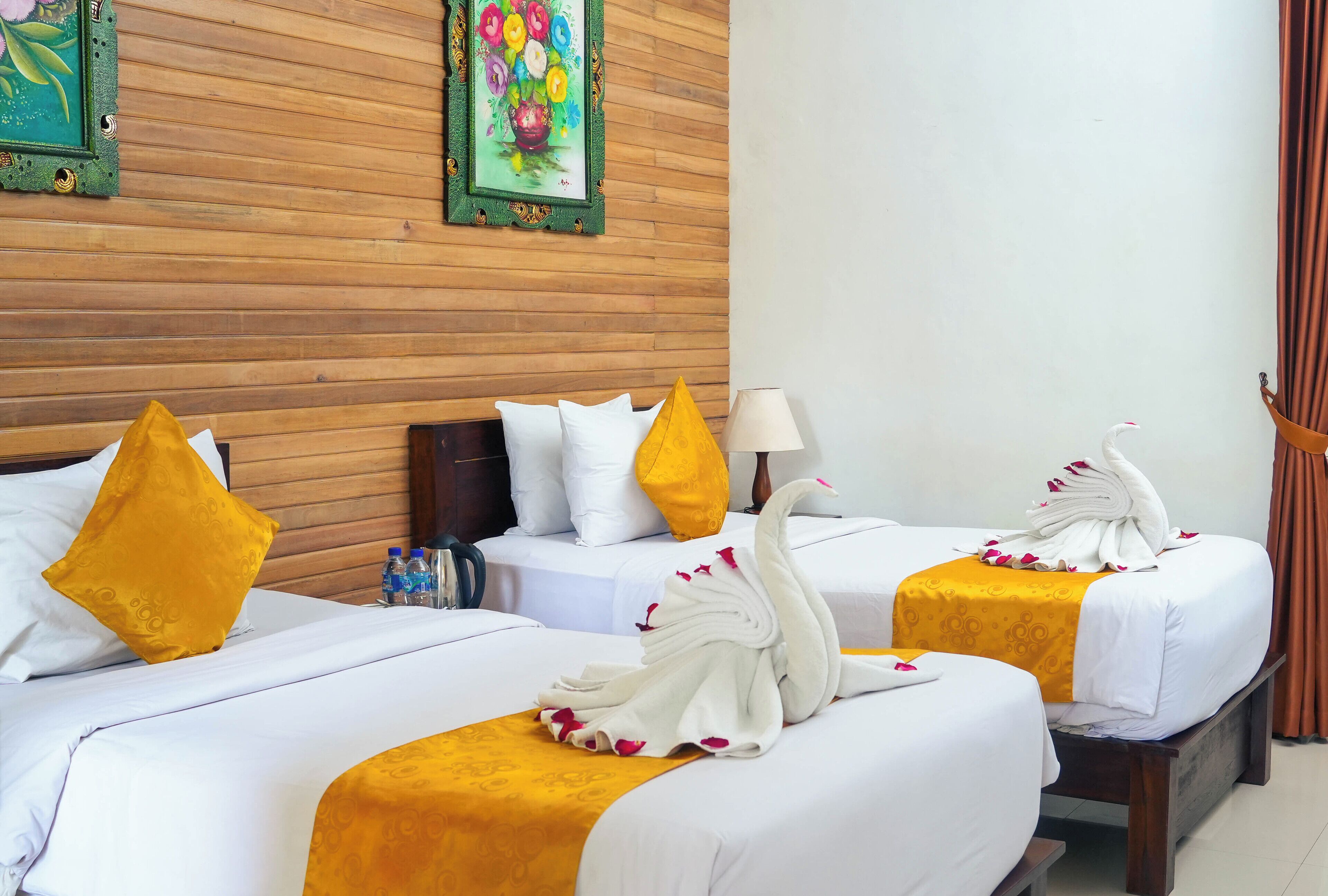 A hotel room with two beds, each with white bedding and a yellow accent. There are decorative swans on the beds, and a painting of flowers on the wall.