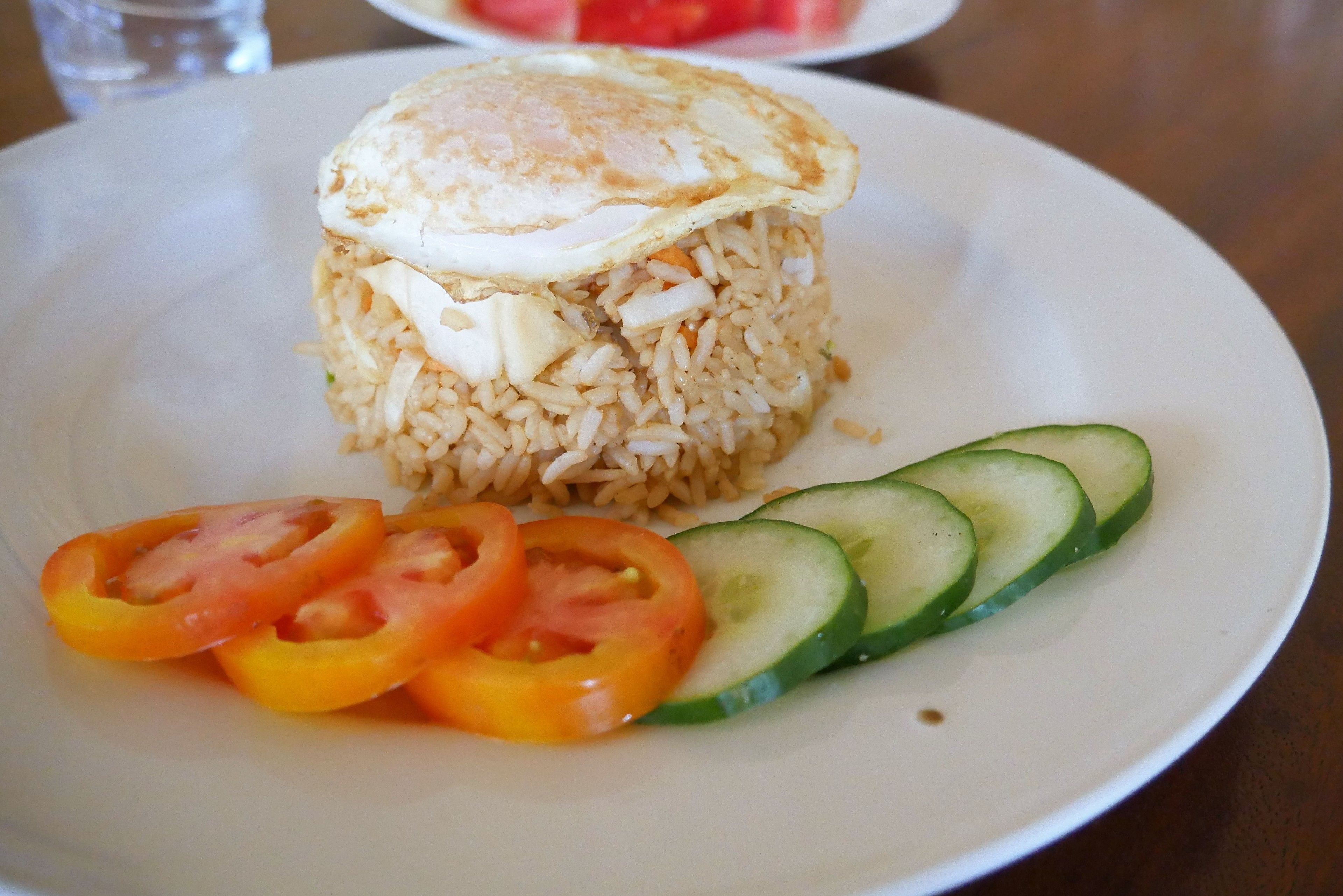 A plate with fried rice topped with a fried egg, accompanied by sliced tomatoes and cucumbers.