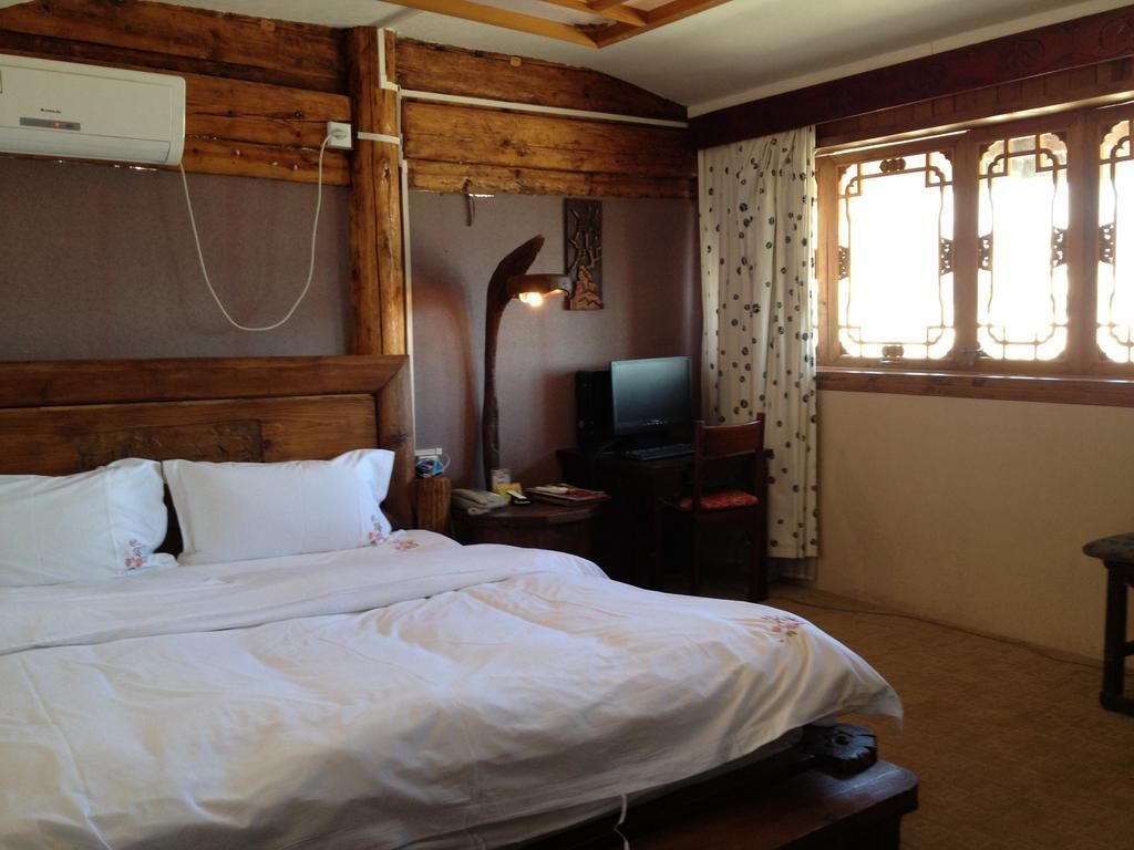 A bedroom with a wooden headboard, a bed with white bedding, a television on a small table, and a window with patterned curtains.