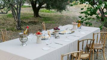 Una mesa colocada al aire libre con mantel blanco, rodeada de sillas de madera y una botella de vino.