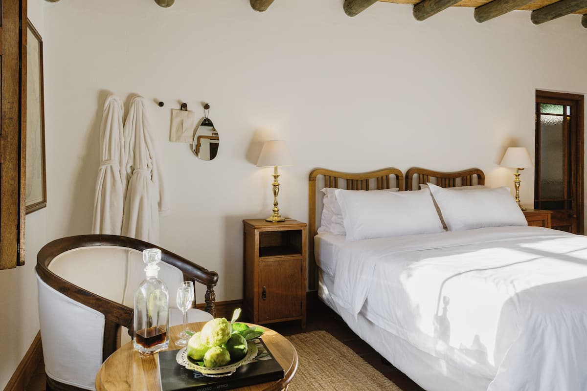 Ein Schlafzimmer mit einem hölzernen Bett, Nachttischlampen, einem Beistelltisch, einem Sessel und einem Tisch mit Flasche und Gläsern.