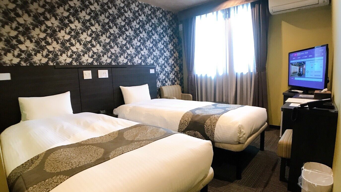 A hotel room with two beds, a desk, a television, and a window with curtains.