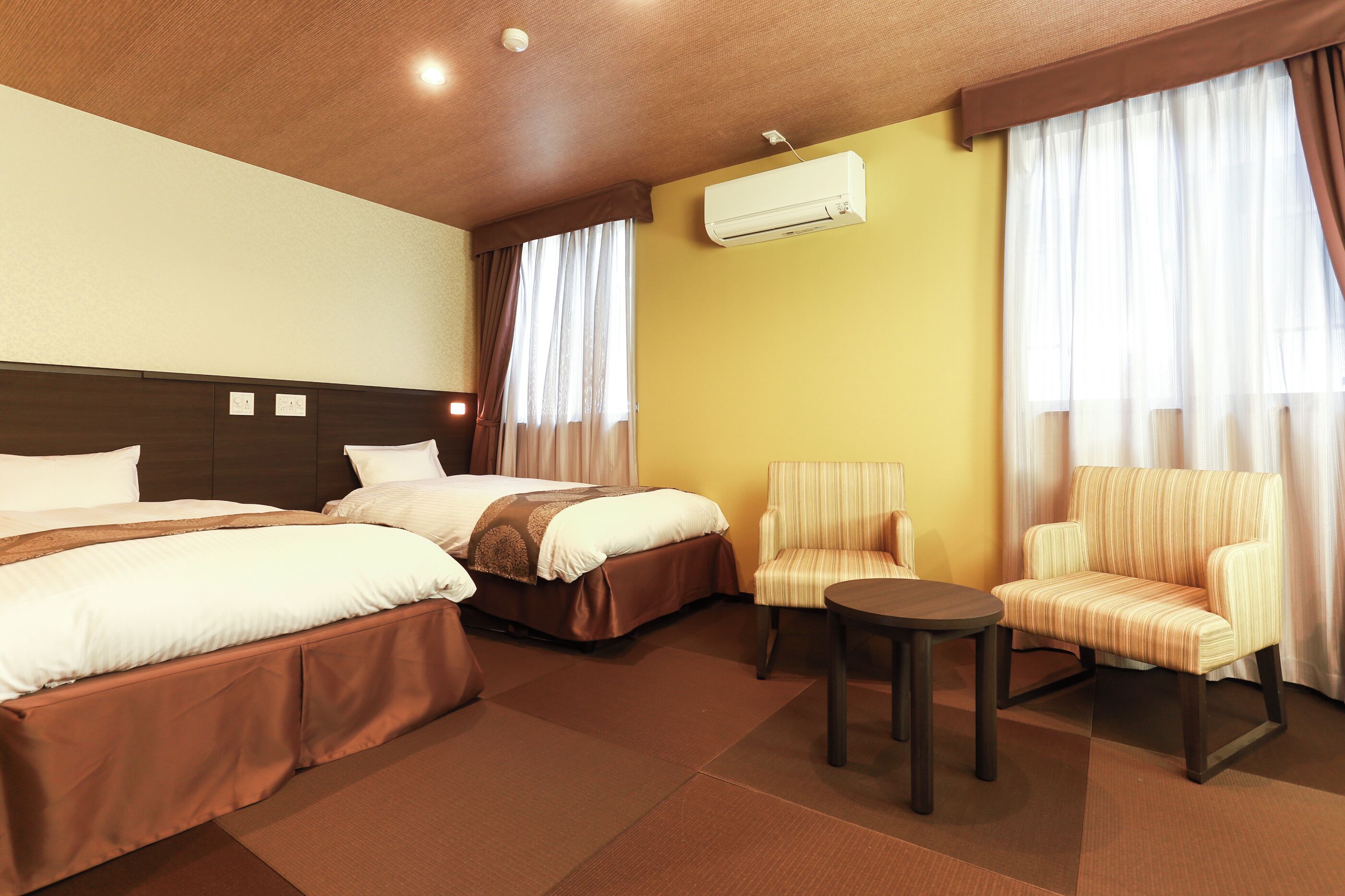 A hotel room with two beds, a small table, two armchairs, an air conditioning unit, and curtains.