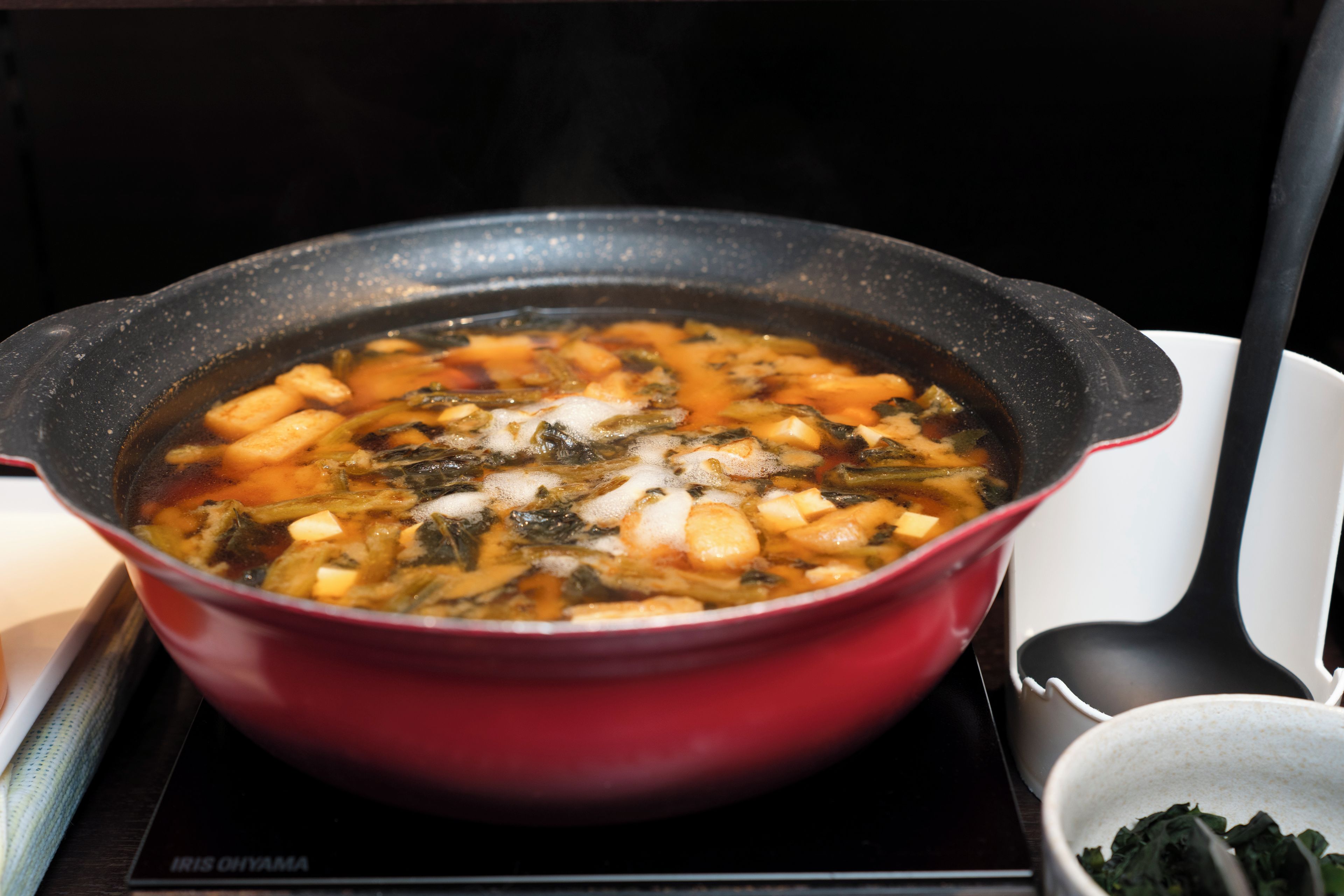 A red pot on a stove with a soup containing vegetables and tofu.