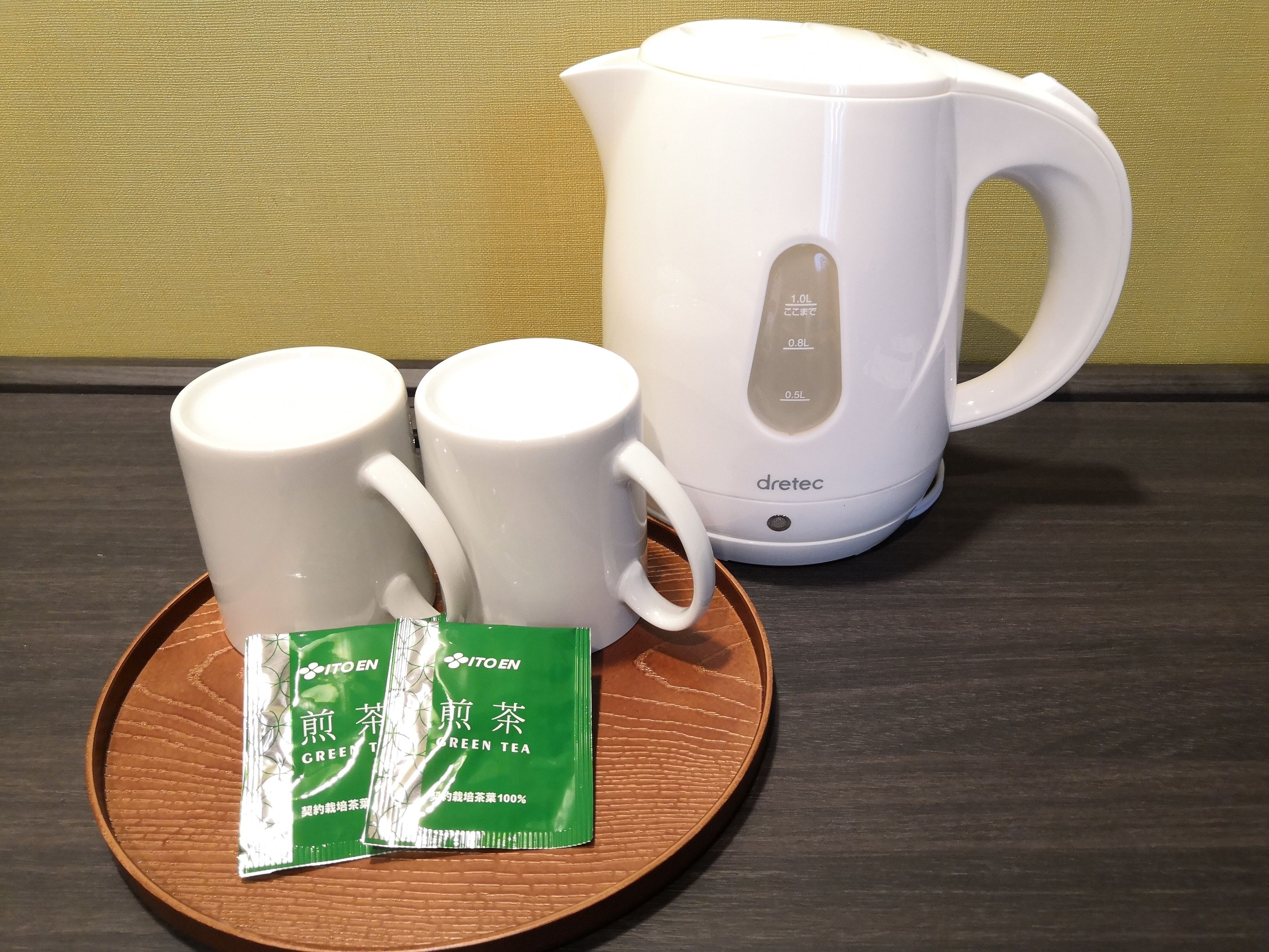 A white electric kettle, two white mugs, and a plate with two green tea packets on a wooden surface.