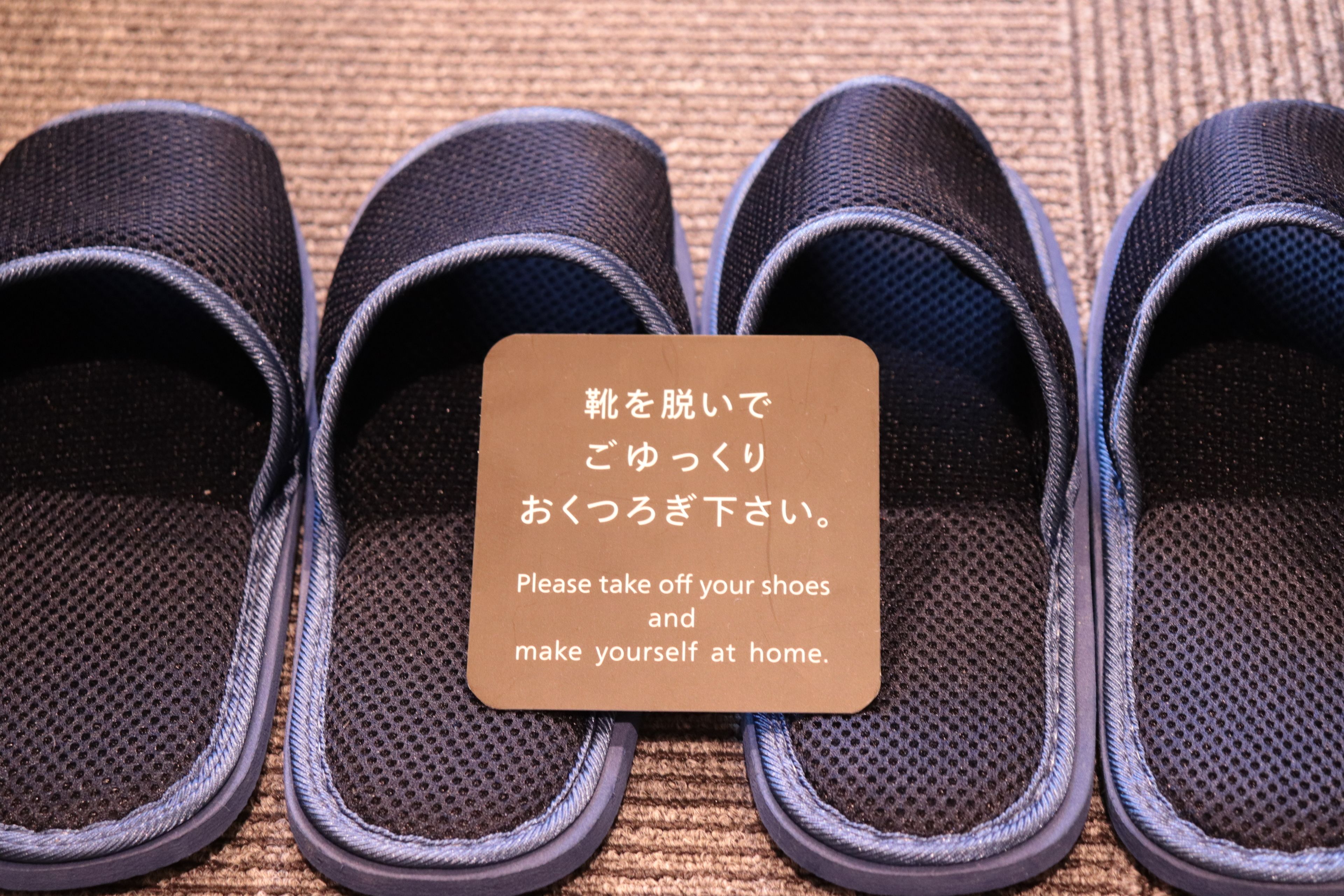 A pair of black slippers with a sign instructing to remove shoes and make oneself at home.