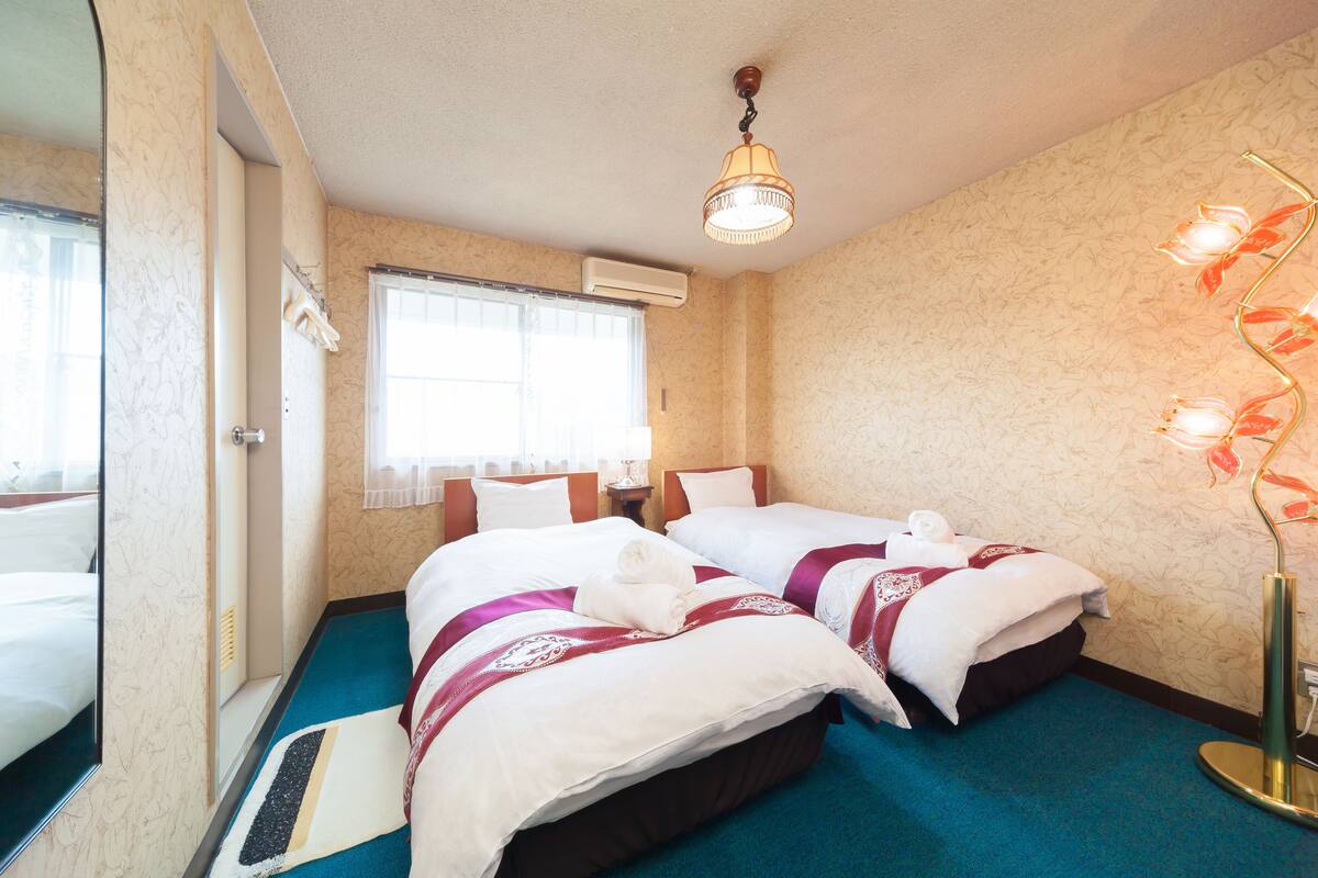 A hotel room with two beds, a blue carpet, a ceiling light fixture, and a window with blinds.