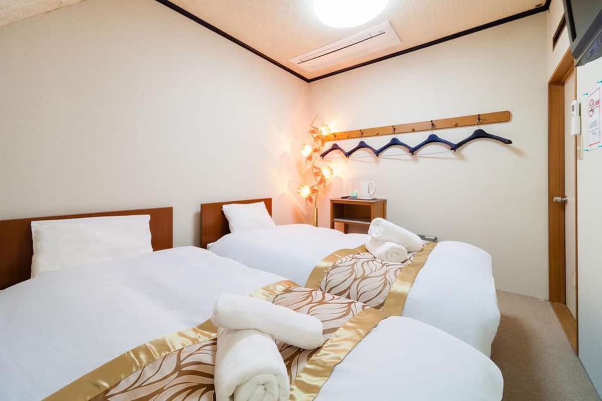 A hotel room with two beds, a bedside table with a lamp, and a wall-mounted towel rack.
