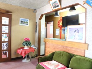 Un área de recepción con un mostrador de madera, un sofá verde, una pequeña mesa con mantel rojo y una planta en maceta.
