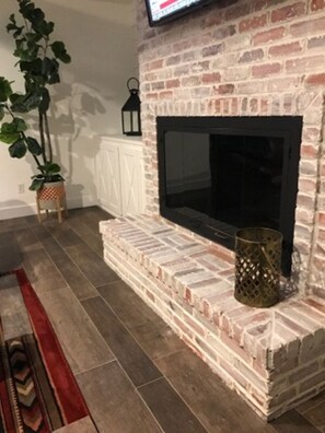 Una sala de estar con chimenea de ladrillo, un televisor de pantalla plana, una planta en maceta y una alfombra estampada.