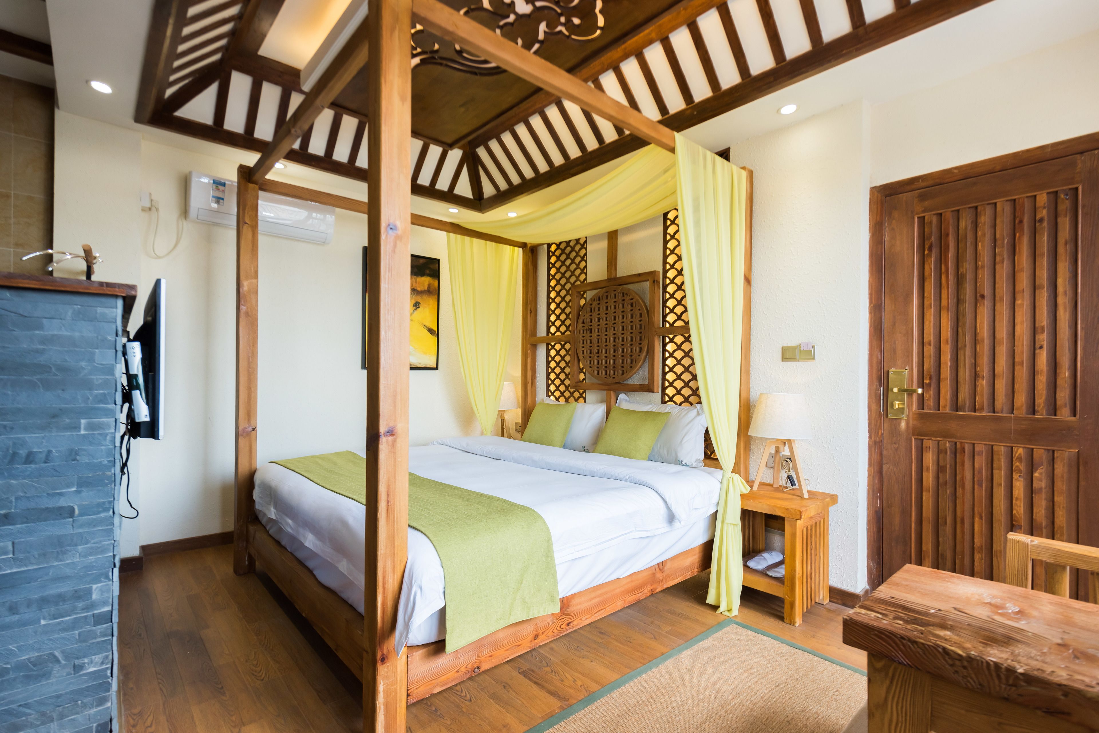 Een hemelbed met houten frame en groen beddengoed, een nachtkastje van hout met een lamp en een houten deur met een verticale panelen decoratie.