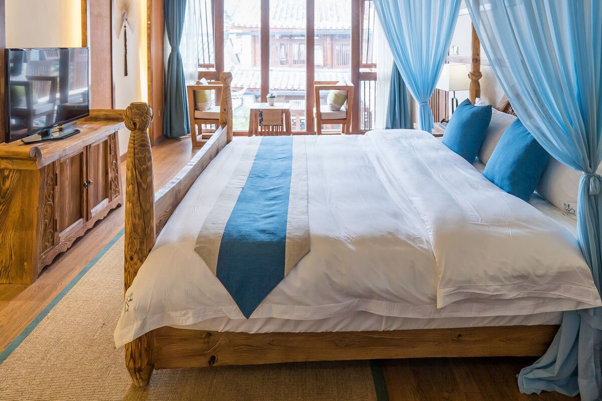 A bedroom with a wooden ceiling, a large bed with blue and white bedding, a television on a wooden stand, and a view of the outside through a window with blue curtains.