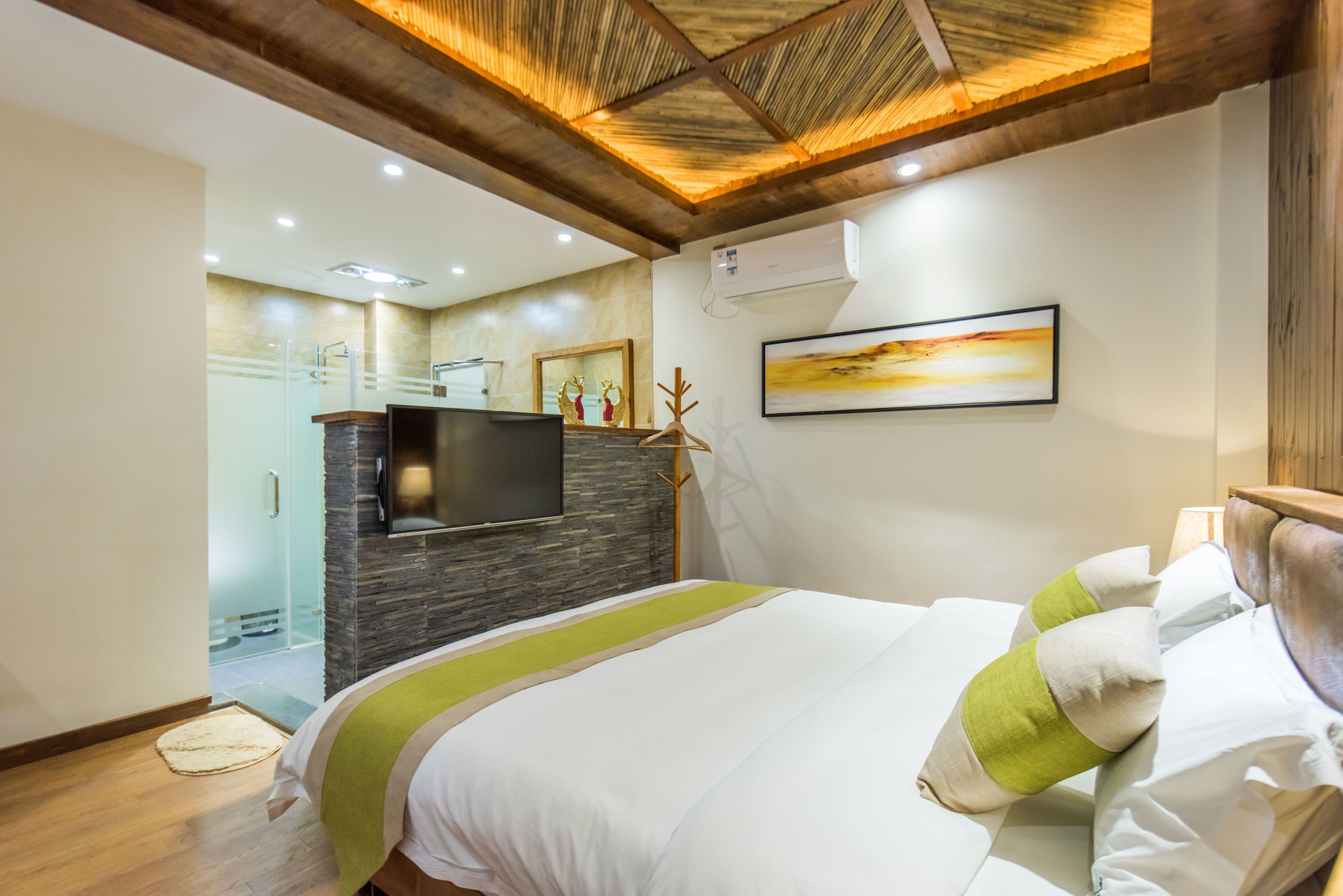 A modern hotel room with a large bed, a flat-screen TV, a stone accent wall, and a ceiling with wooden beams.