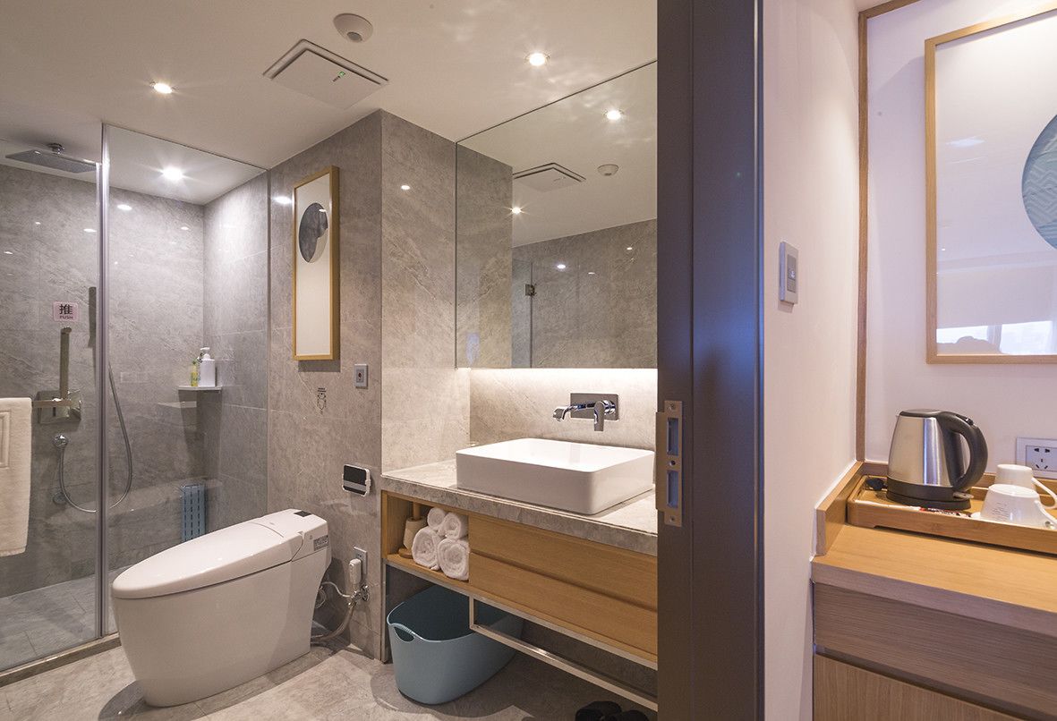 A modern bathroom with a glass shower enclosure, a white toilet, a sink with a mirror above, and a wooden vanity with a kettle and cups.