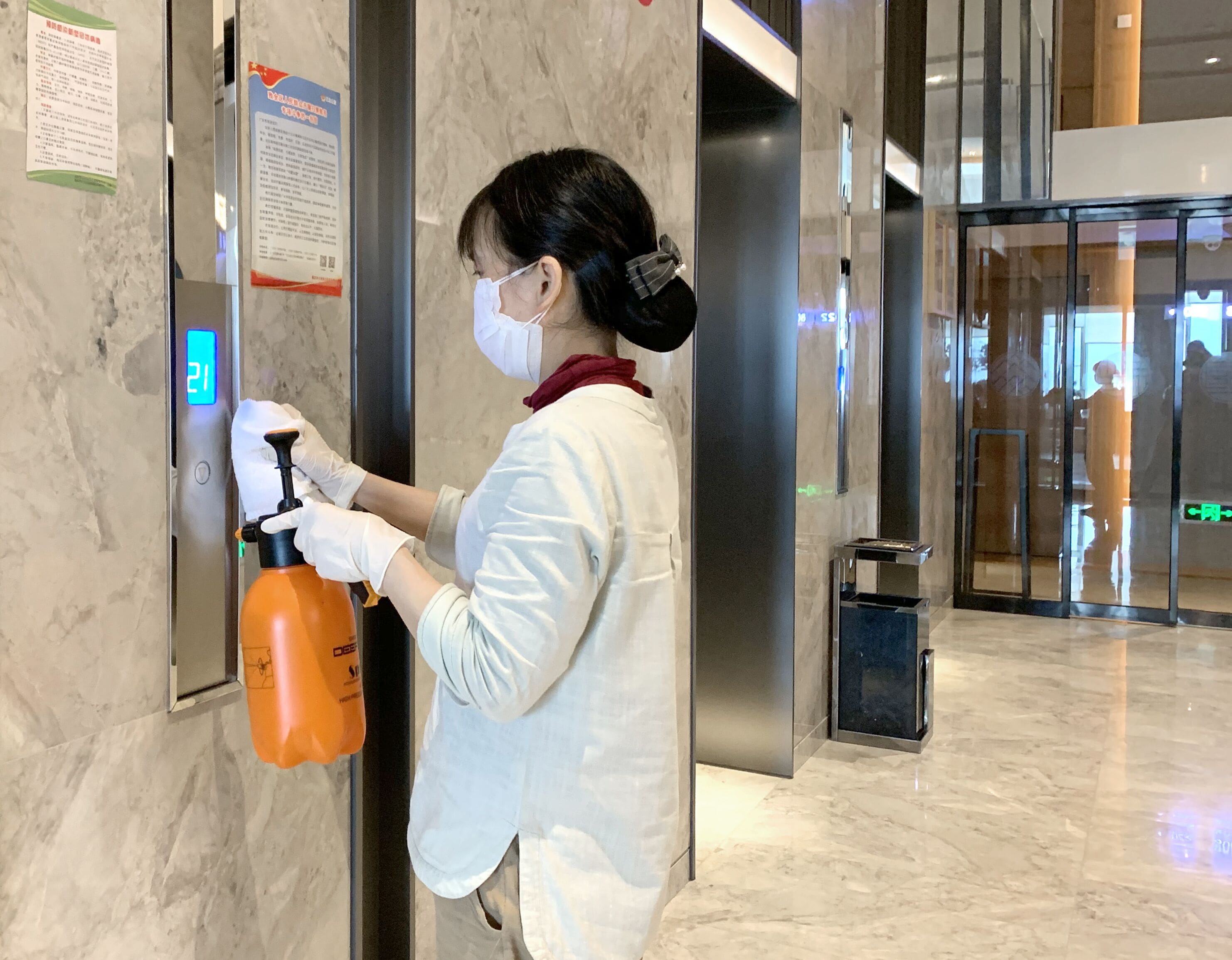 A person using a hand sanitizer dispenser in a hallway with marble flooring and glass doors in the background.