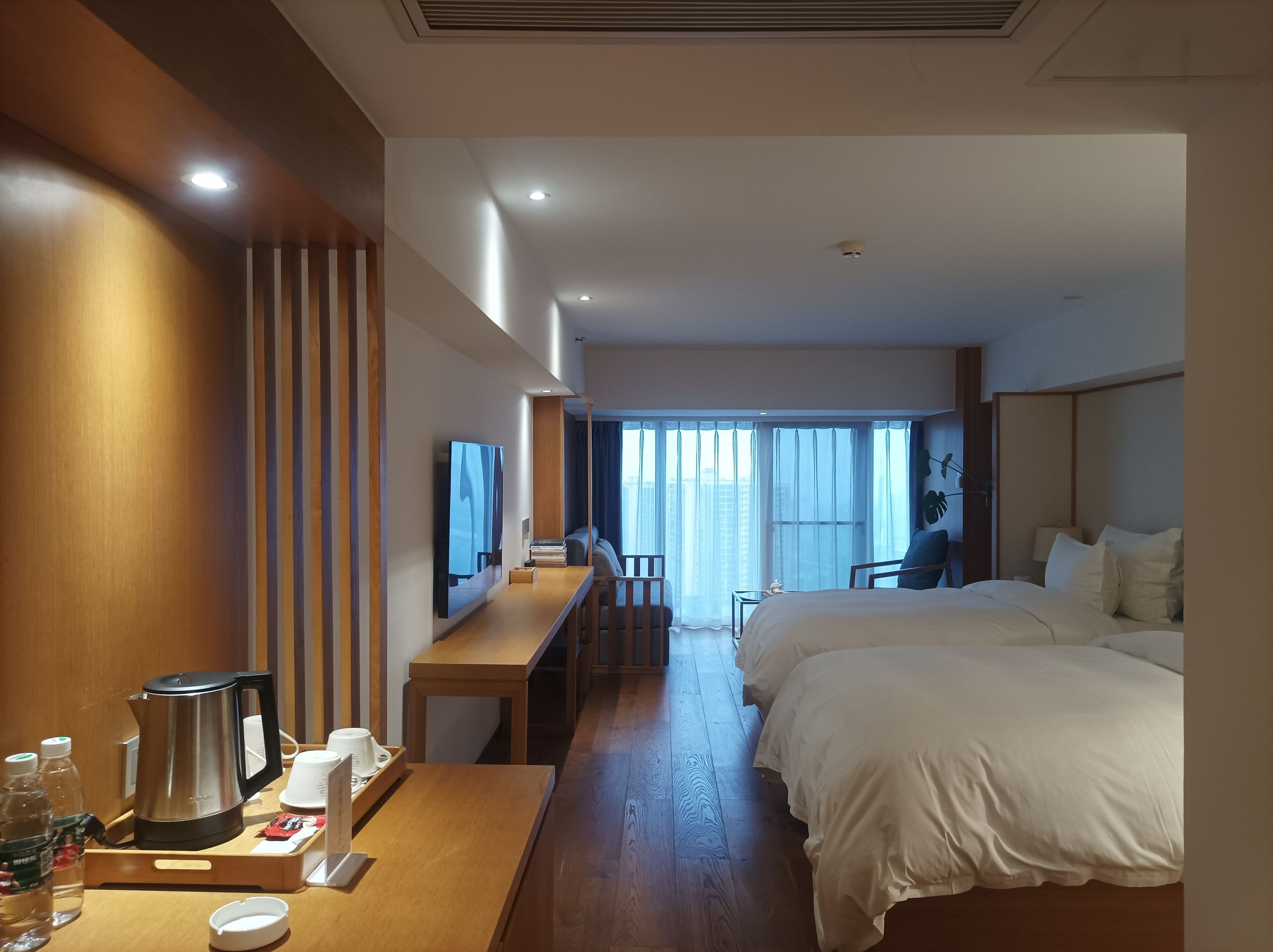 Ein Hotelzimmer mit Holztisch, Bett, Fernseher und Blick auf die Stadt durch große Fenster.