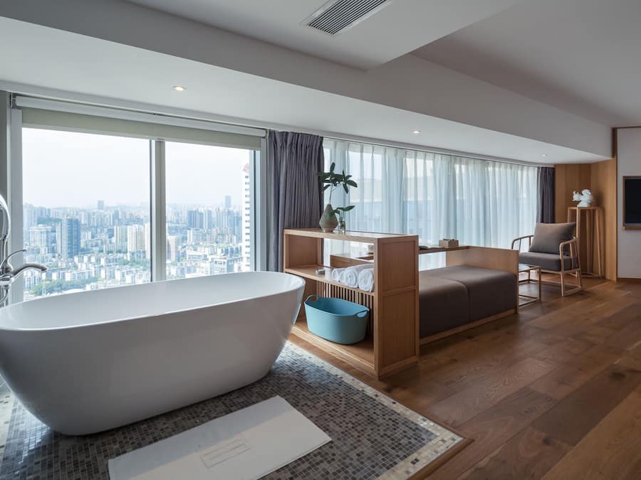Un bagno moderno con una vasca freestanding, un'ampia finestra vista città e un'area relax con divano e poltrone.
