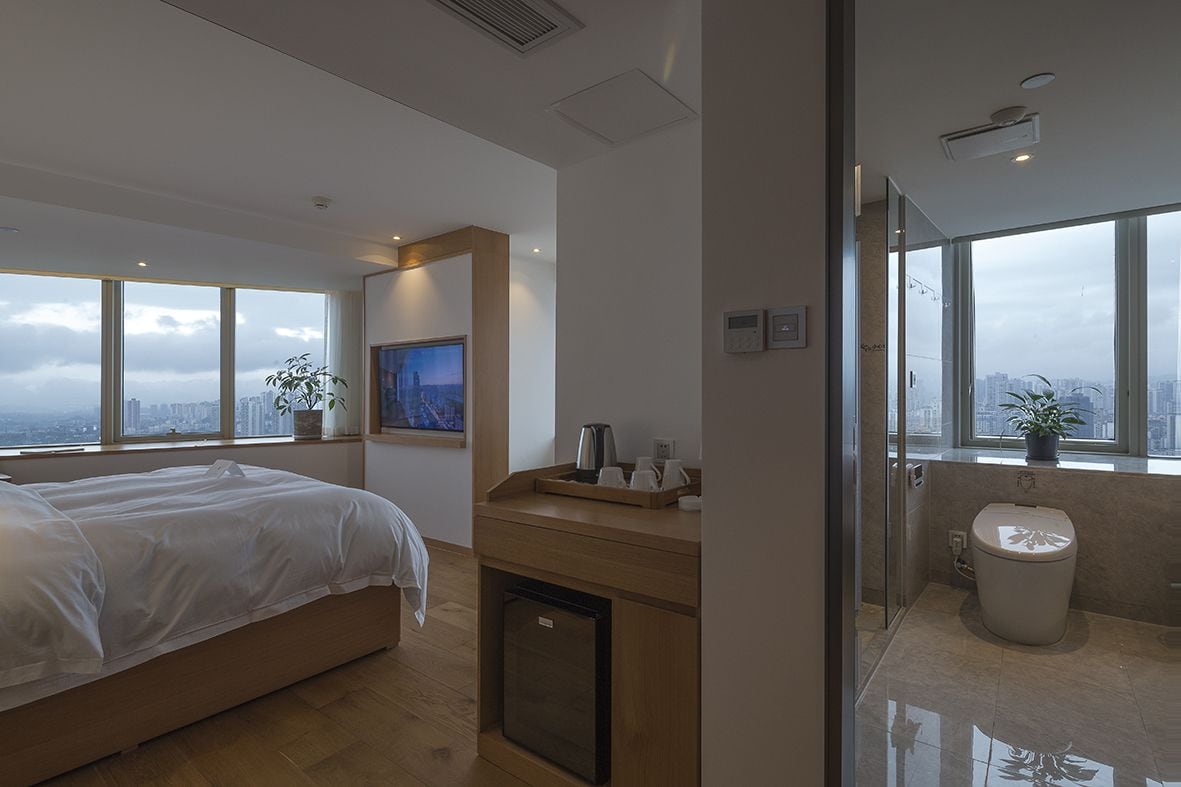 Una habitación de hotel moderna con una cama grande, una kitchenette pequeña, un inodoro y un ventanal con vistas a la ciudad.