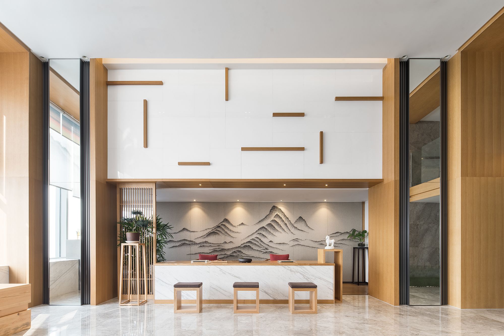 Un área de recepción moderna con un mostrador de madera, tres taburetes y una pared decorativa con un paisaje de montaña.
