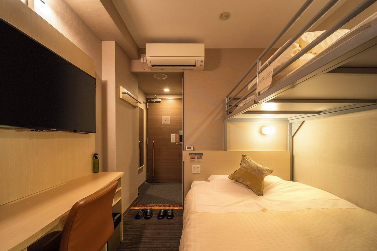 Habitación de hotel compacta con una cama litera, un escritorio con silla, un televisor de pantalla plana y un zapatero.