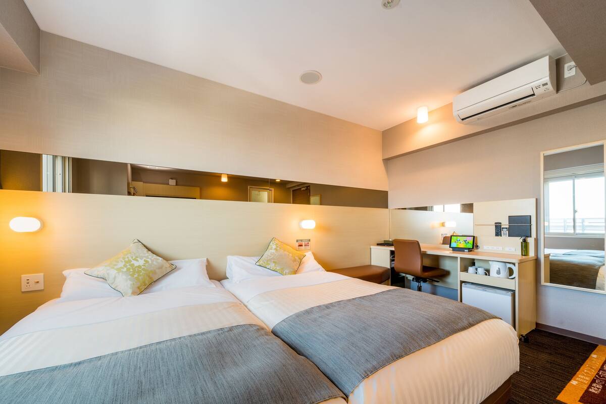 Habitación de hotel con dos camas, un escritorio, una silla, un espejo y un equipo de aire acondicionado.