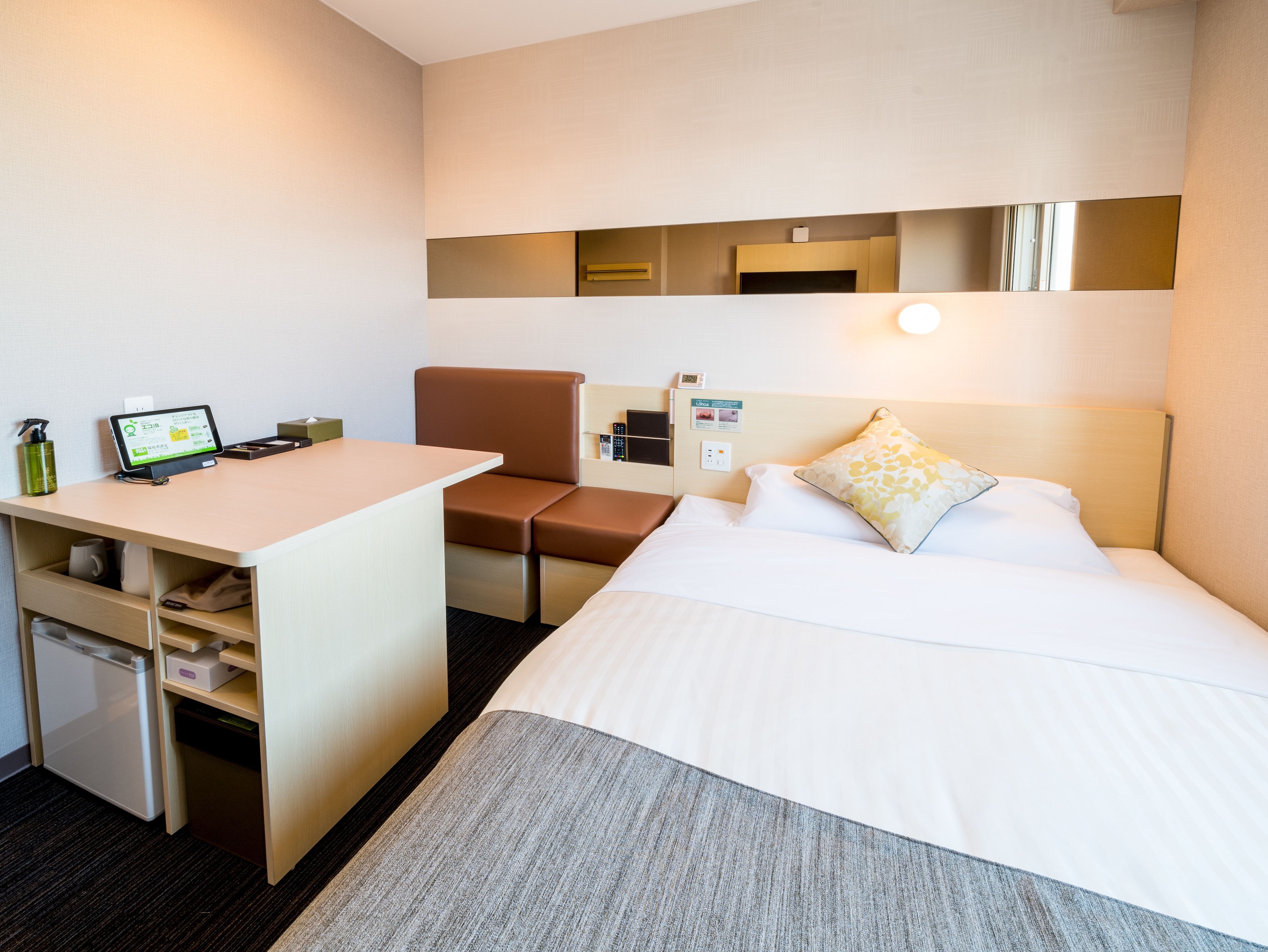 A hotel room with a bed, a desk, a chair, a small refrigerator, and a bedside table.