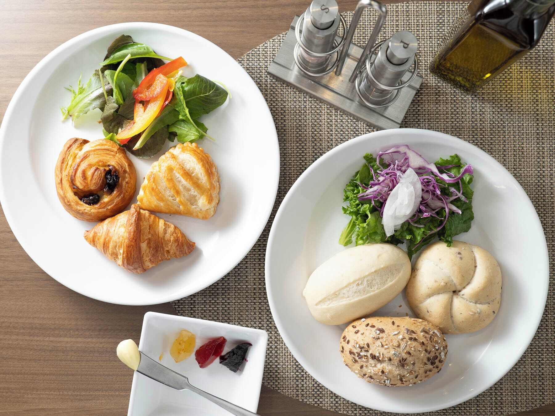 Deux assiettes blanches contenant divers aliments, notamment des croissants, des petits pains et une salade au chou rouge, sont posées sur une table en bois recouverte d’une nappe tressée.