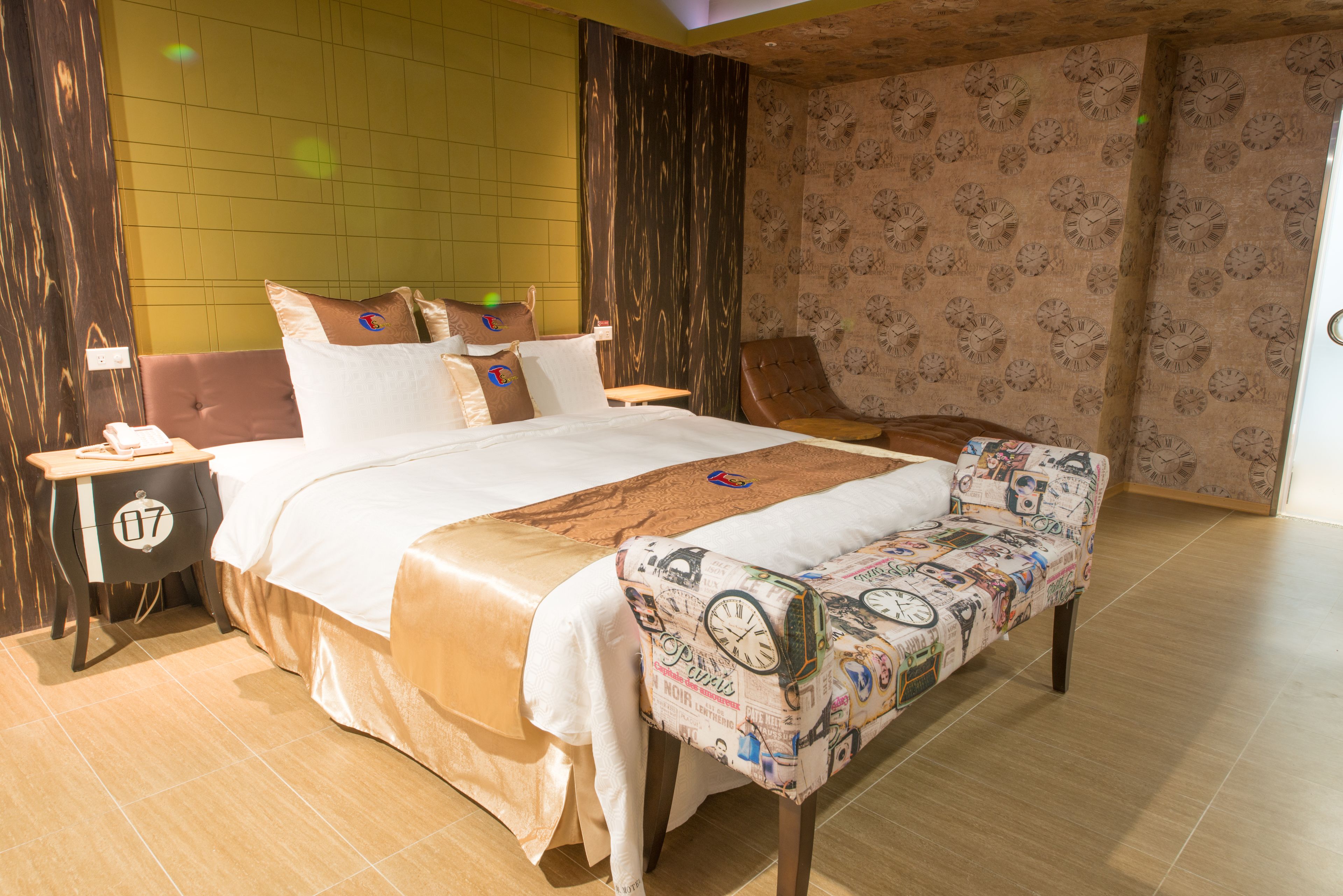 A well-appointed bedroom with a large bed, a bedside table, a nightstand, a bench, and a wall with a clock pattern.