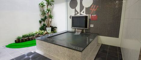 Un moderno espacio de spa con un jacuzzi de vidrio, televisor y un mural de mosaicos.