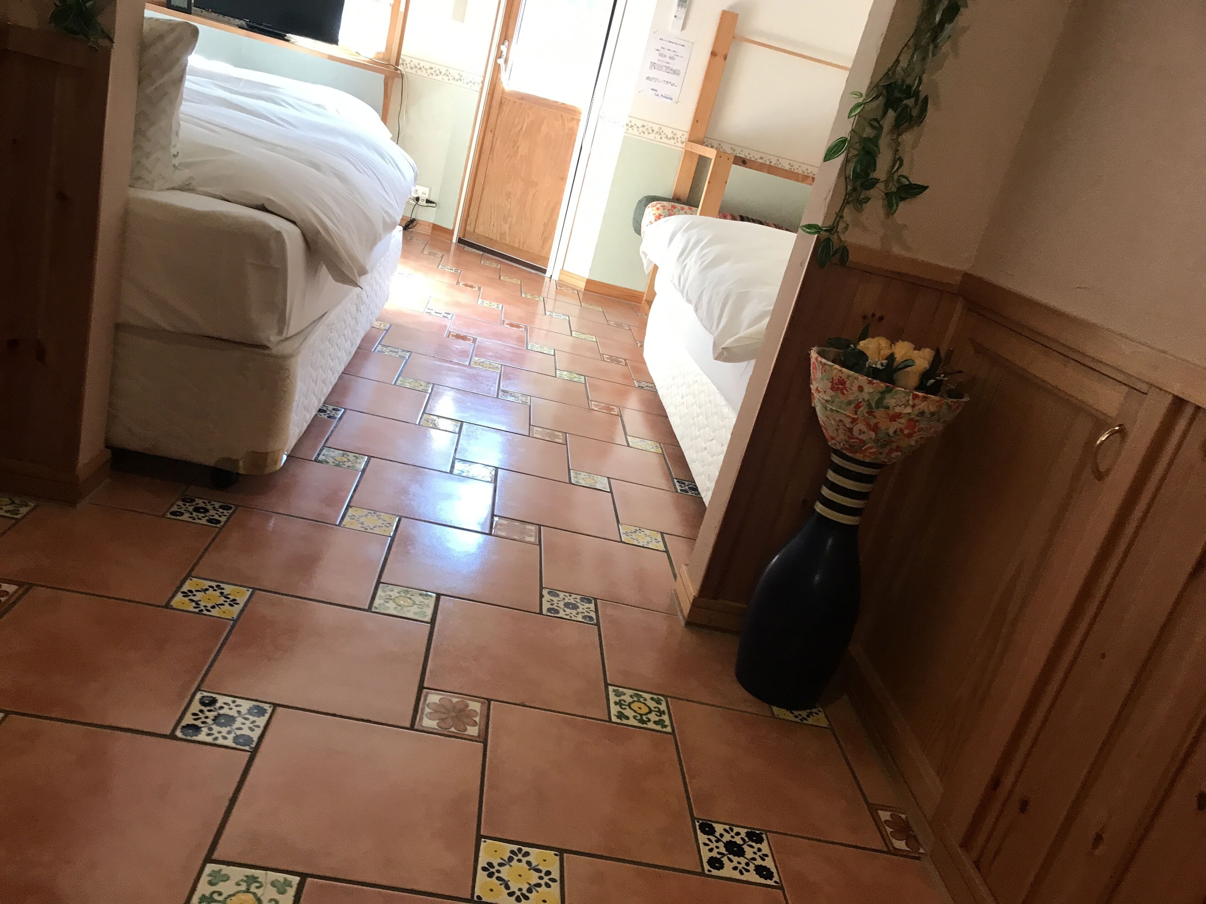 A hotel room with two beds, a tiled floor, a wooden door, and a vase with flowers.