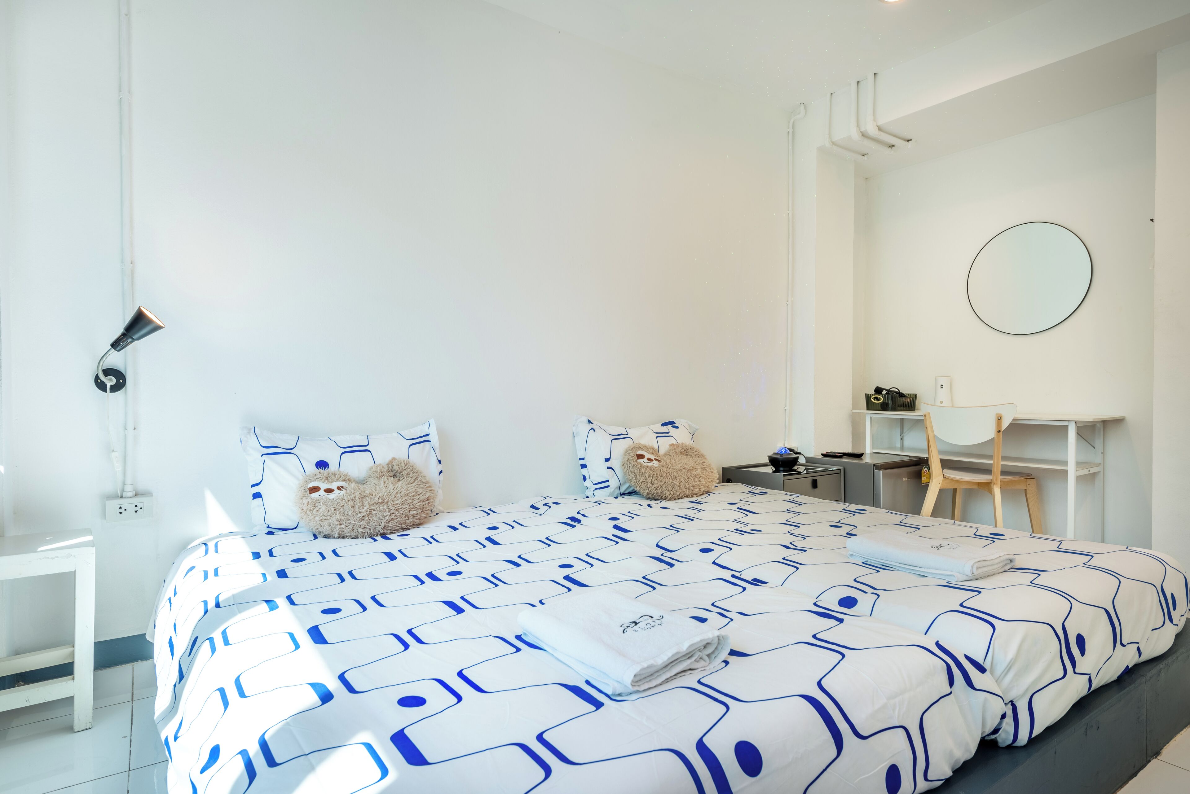 Een net opgemaakt bed met een bedspreide met een geometrisch patroon, twee teddyberen op de kussens, een bureau met een stoel en een ronde spiegel aan de muur.