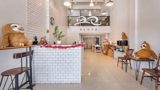 A cozy cafe with a sloth-themed decor, featuring a sloth statue at the counter and on the wall, a small plant, and a sign that reads "SLOTH". - Sloth Hostel Don Mueang (Bangkok)