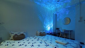 A bedroom with a patterned bedspread, a bedside table with a lamp, a chair, and a desk with a chair. - Sloth Hostel Don Mueang (Bangkok)
