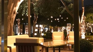 Zona salotto all'aperto con tavoli rotondi e sedie imbottite, circondata da alberi e illuminata da luci decorative.