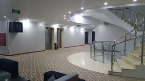 Et hotellobby med en buet trappe, tæppede gulve og en receptionsdisk.