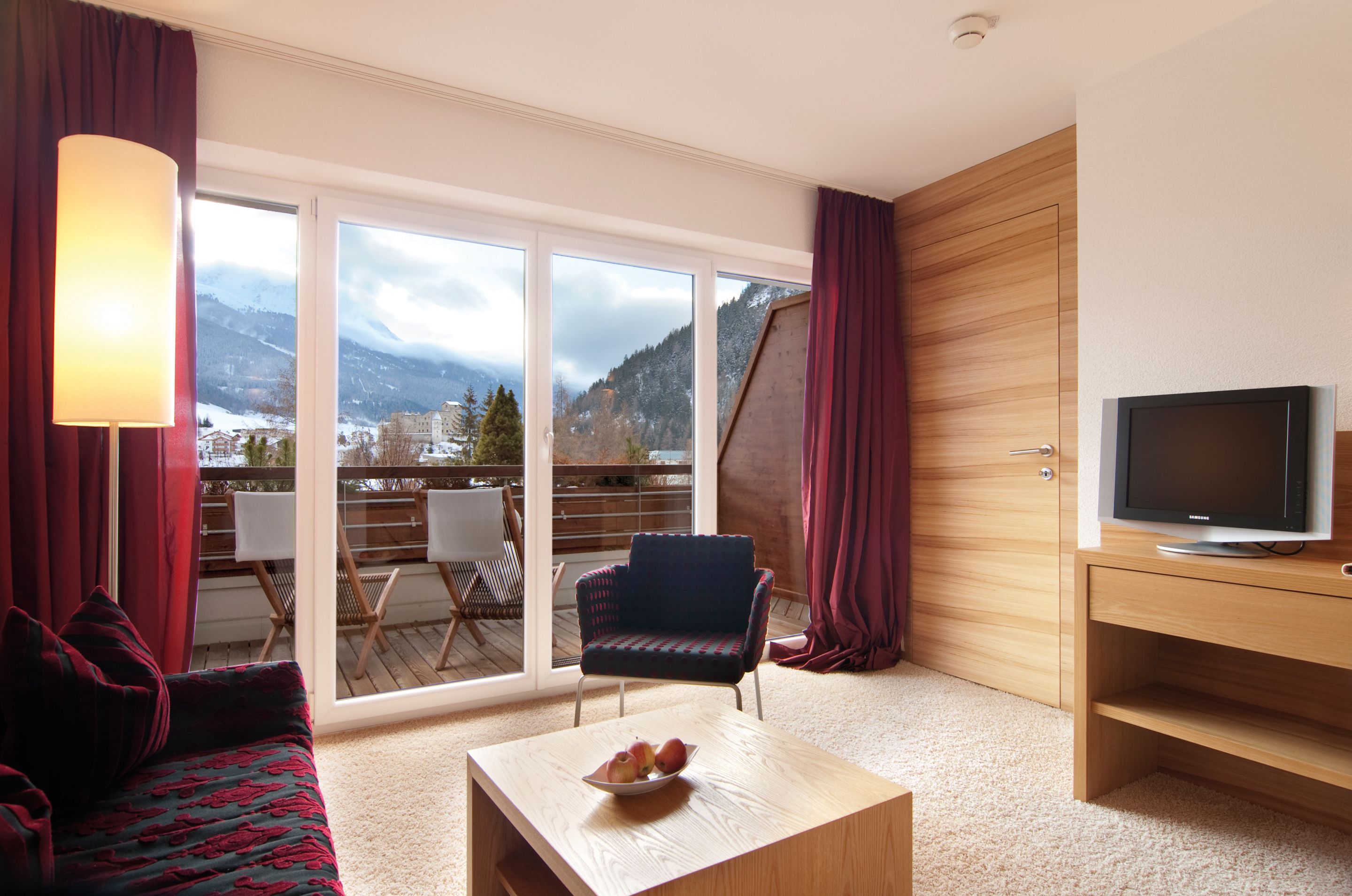 Quarto de hotel com sofá, mesa com frutas, televisão, varanda com cadeiras e vista para as montanhas.